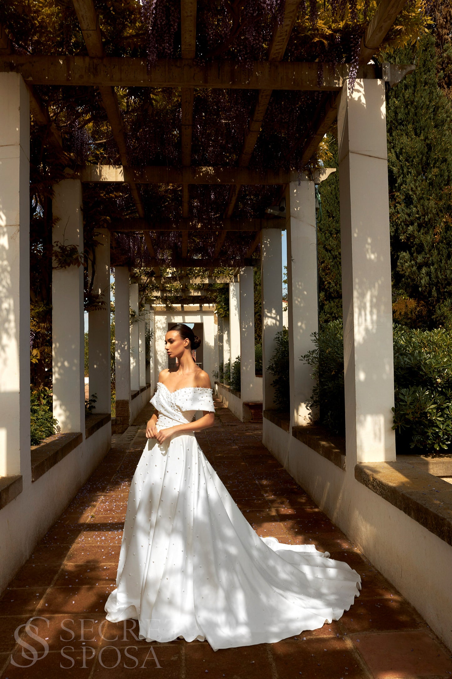 Bride in plain Mikado A-line wedding gown with pearl details, off-the-shoulder crossed bodice, chapel train, and zip closure.