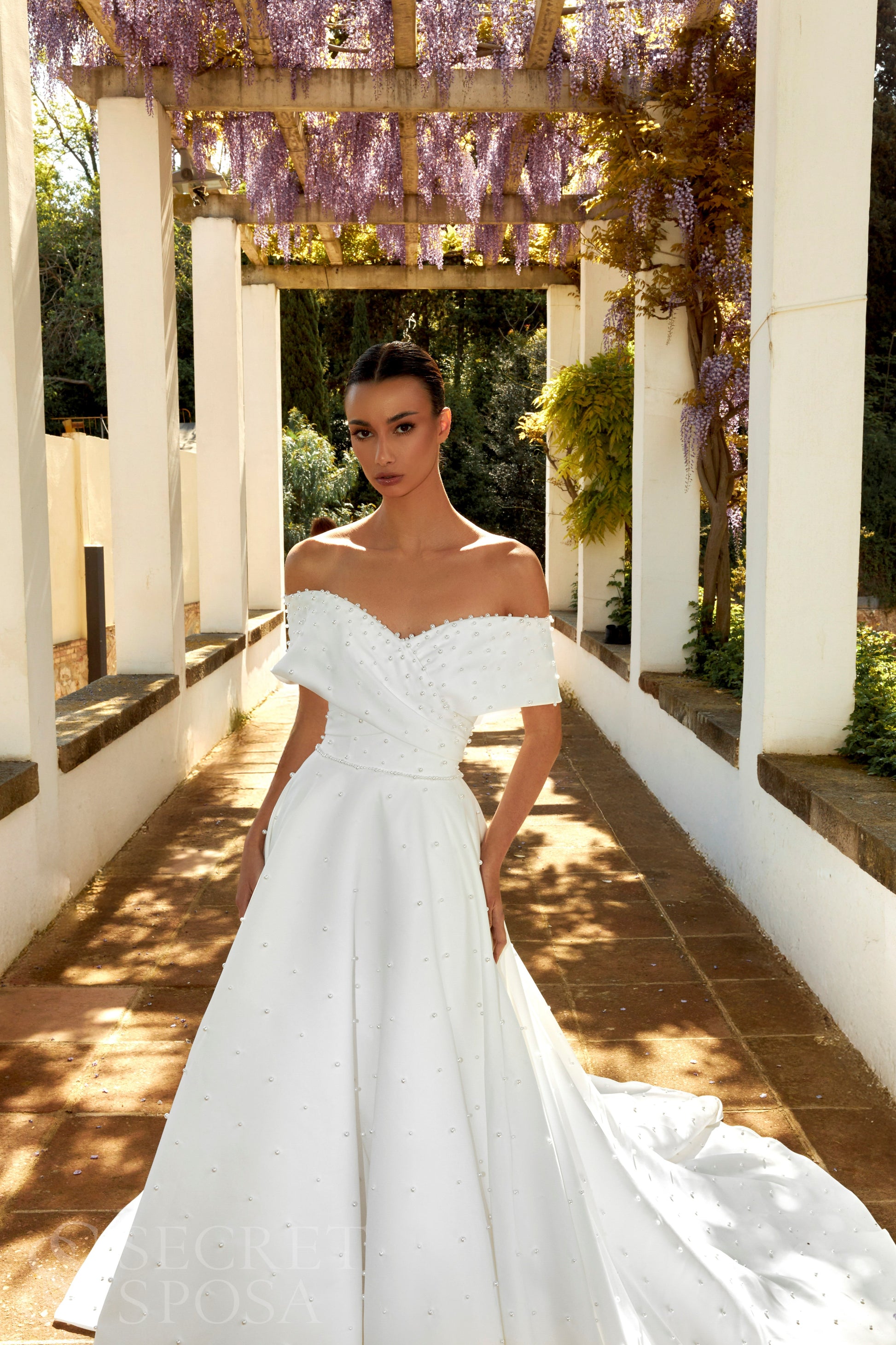 Bride in plain Mikado A-line wedding gown with pearl details, off-the-shoulder crossed bodice, chapel train, and zip closure.