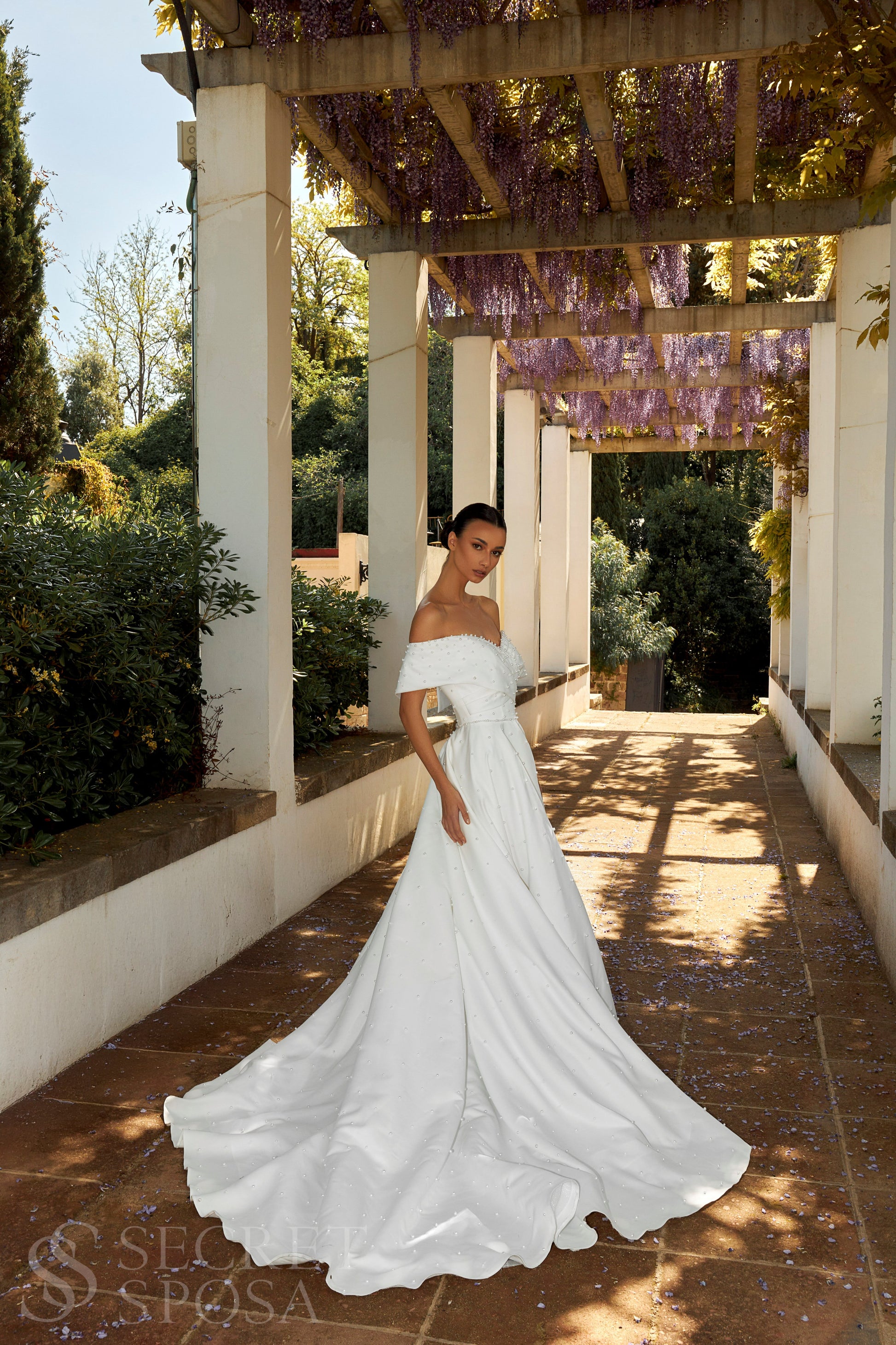 Bride in plain Mikado A-line wedding gown with pearl details, off-the-shoulder crossed bodice, chapel train, and zip closure.