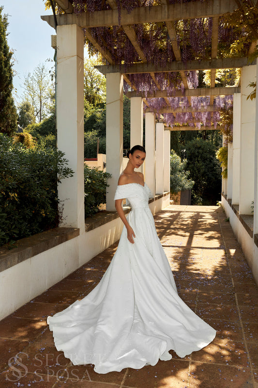 Bride in plain Mikado A-line wedding gown with pearl details, off-the-shoulder crossed bodice, chapel train, and zip closure.