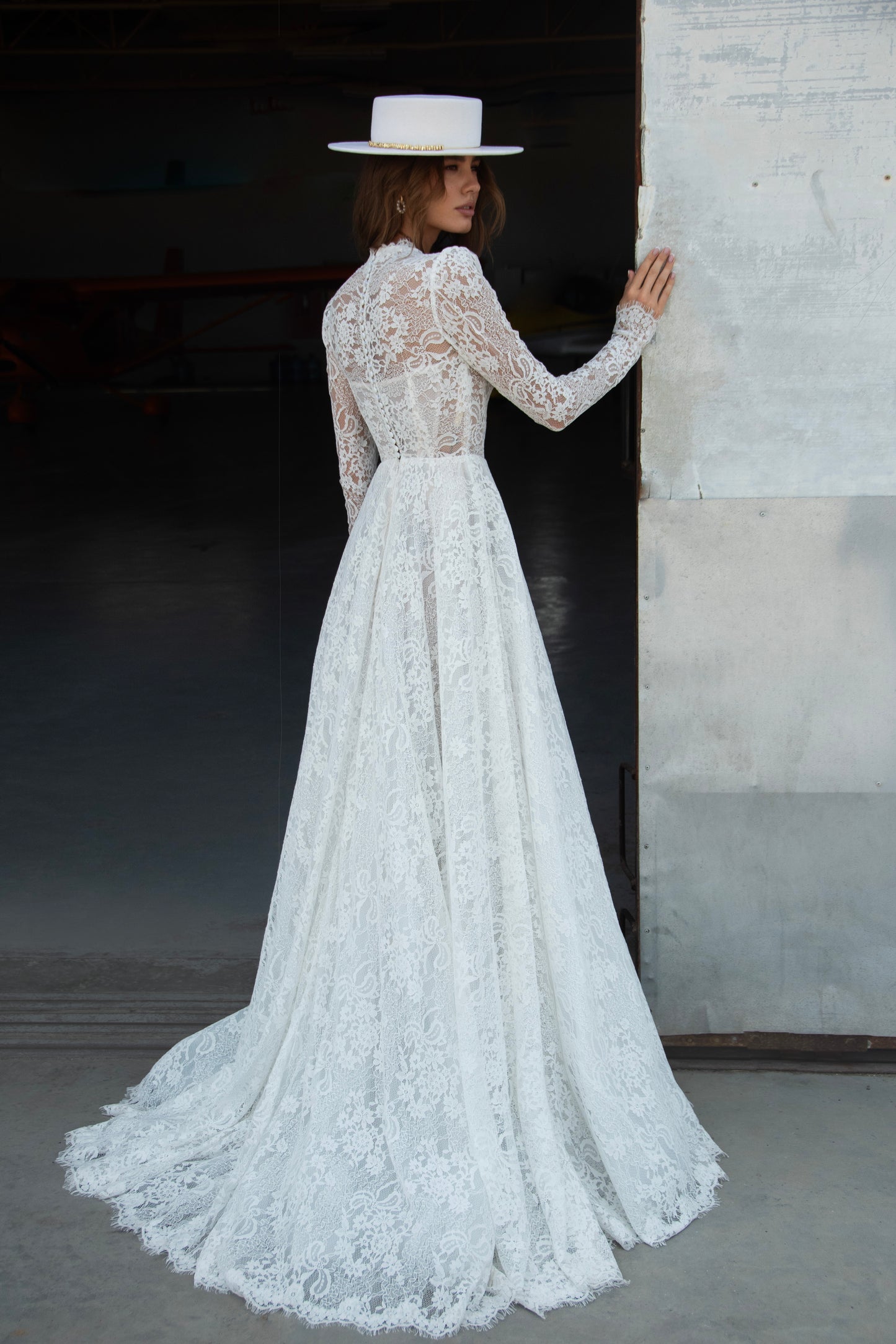 A-line lace wedding dress with long sleeves, V-neckline, high buttoned back, and flowing court-length train featuring detailed lace texture and modern bridal design