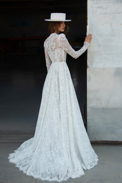 A-line lace wedding dress with long sleeves, V-neckline, high buttoned back, and flowing court-length train featuring detailed lace texture and modern bridal design