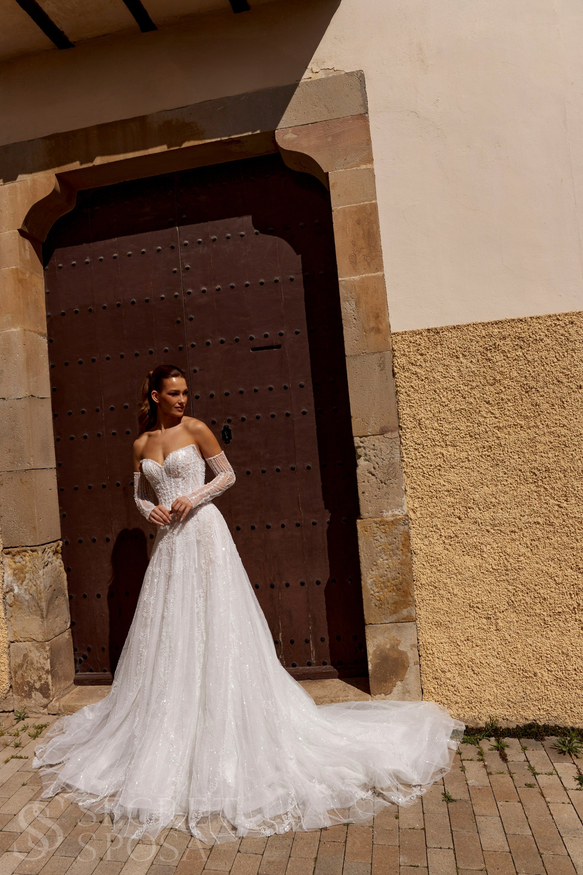 Bride in A-line tulle wedding dress with pearl and sequin embroidery, V-neckline, removable long off-the-shoulder sleeves, straight open back, and chapel train.