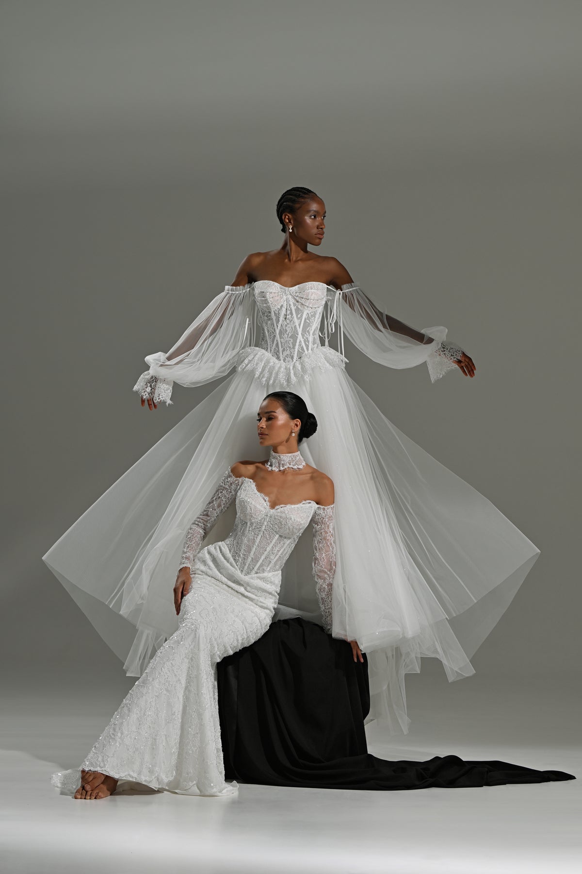 Lace and tulle ball gown wedding dress with corset bodice and peplum waist – two brides, one standing in the tulle overskirt and one seated on a black block