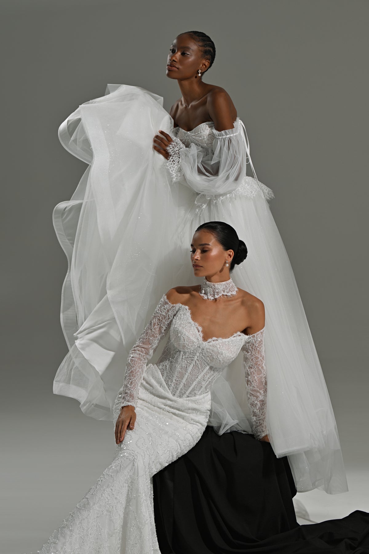 Lace mermaid wedding dress with sweetheart corset and tulle overskirt – two brides seated on a black block
