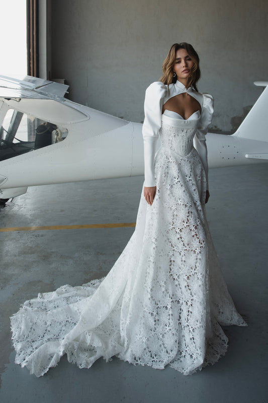 Bride in lace A-line wedding dress with satin bolero, sweetheart corset bodice, long sleeves, and chapel train.