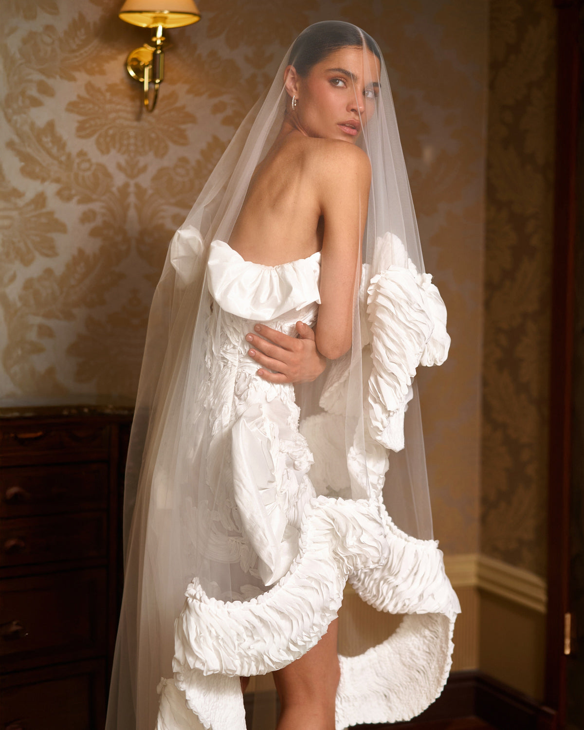 Mini wedding dress seen from behind with a low lace-up back and a flowing sheer cape-veil with ruffled edges.