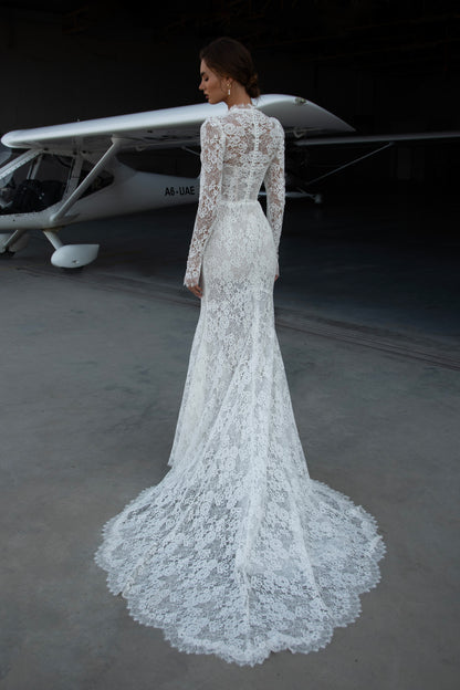 Bride wearing long-sleeve lace fit-and-flare wedding dress with scalloped neckline and chapel train, photographed beside an airplane hangar.