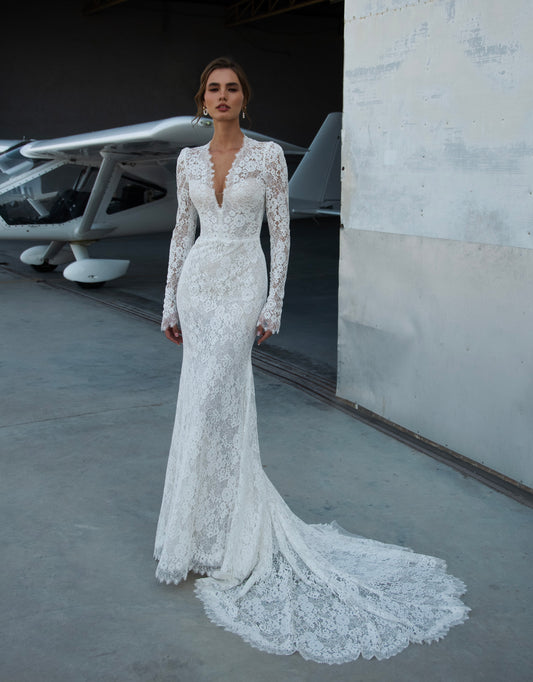 Bride wearing long-sleeve lace fit-and-flare wedding dress with scalloped neckline and chapel train, photographed beside an airplane hangar.