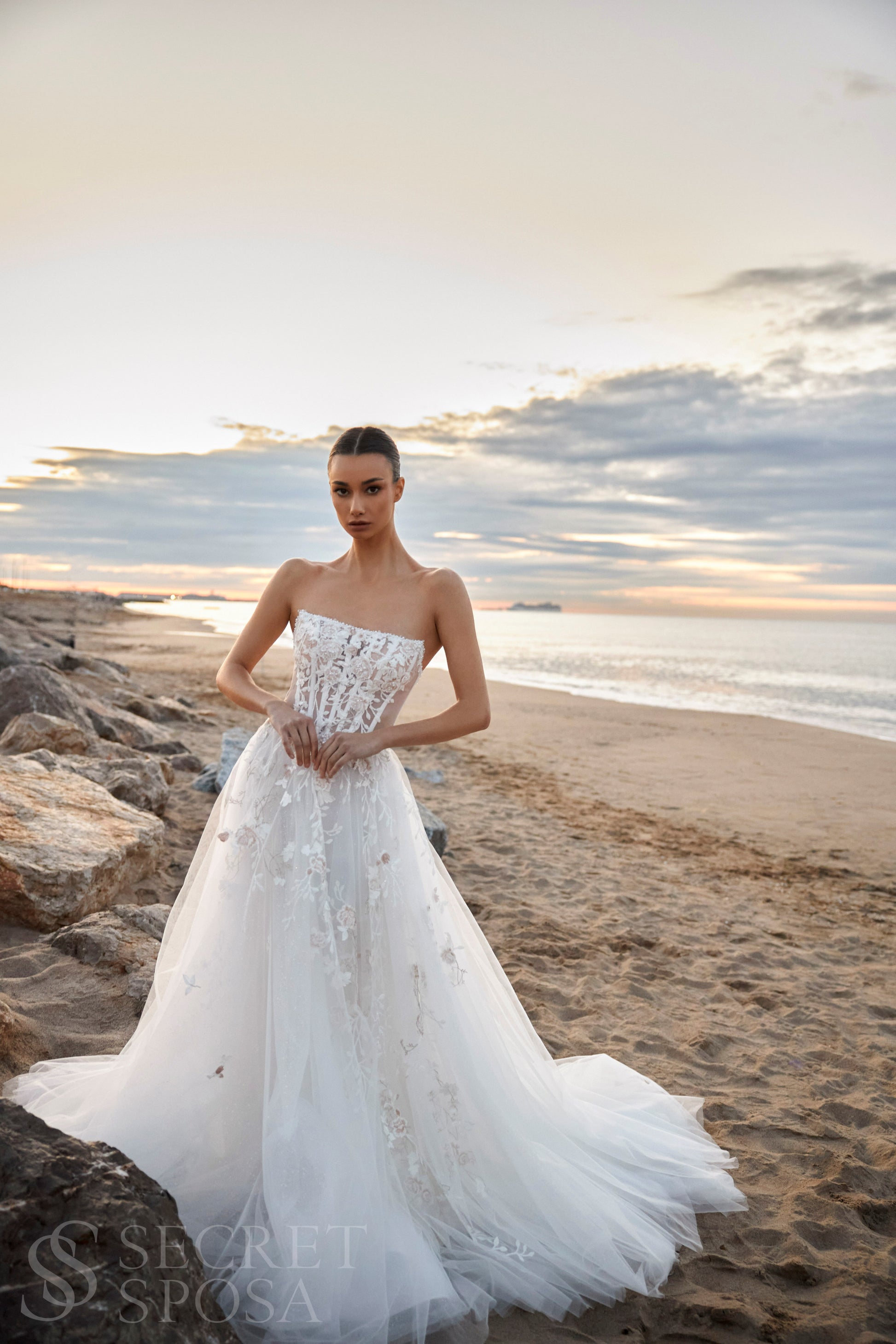 Bride in strapless floral lace and tulle A-line wedding gown with straight neckline, drop-down waist, and chapel train.