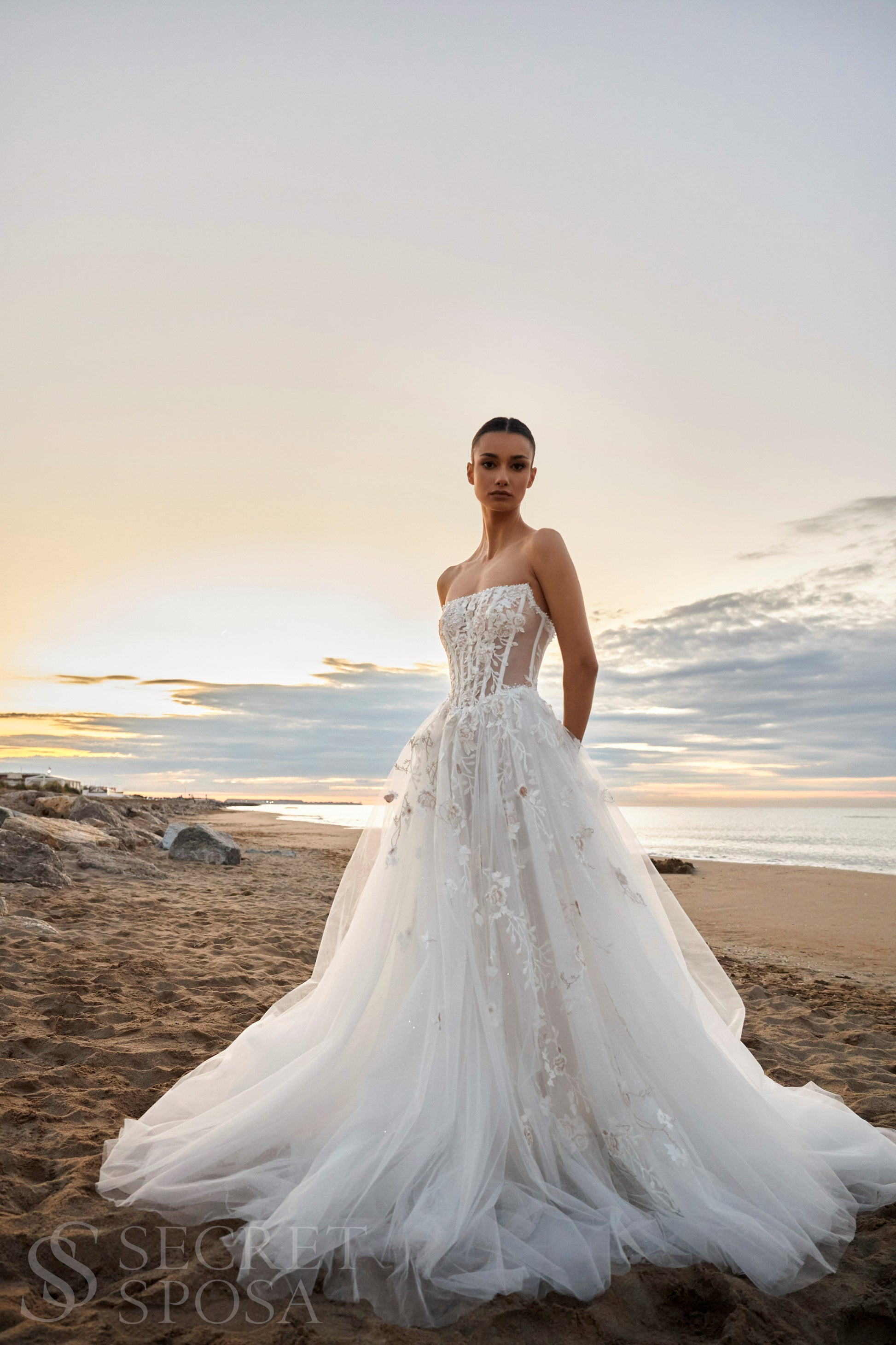 Bride in strapless floral lace and tulle A-line wedding gown with straight neckline, drop-down waist, and chapel train.