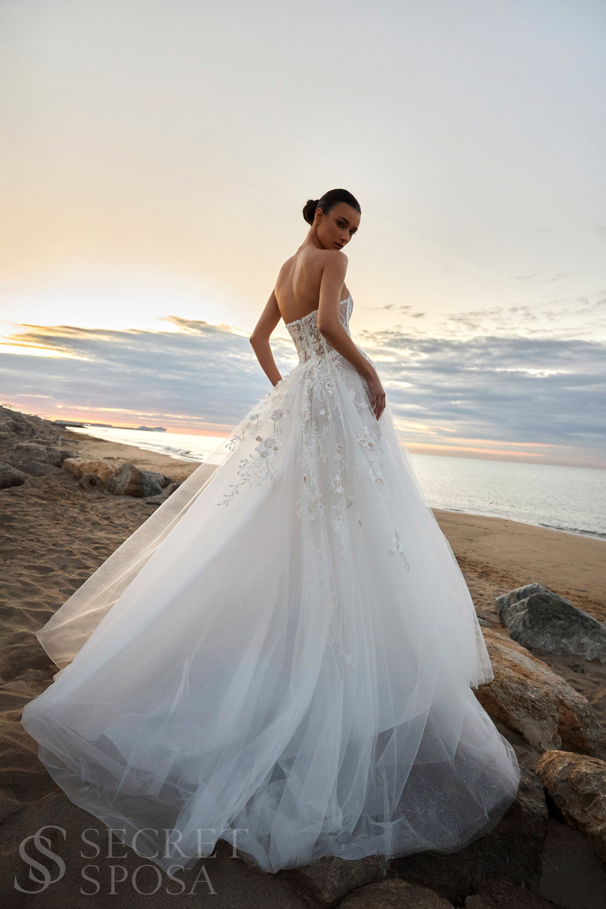 Bride in strapless floral lace and tulle A-line wedding gown with straight neckline, drop-down waist, and chapel train.