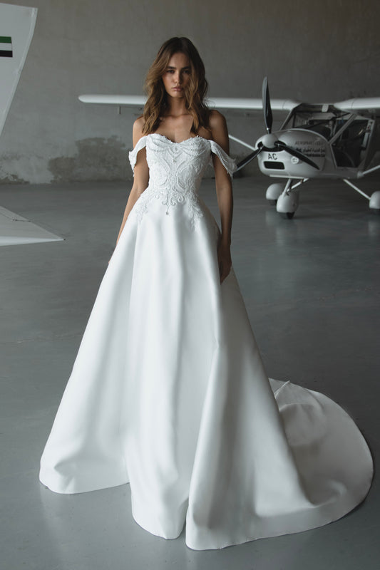 Bride wearing off-the-shoulder Mikado A-line wedding gown with beaded lace bodice and flowing skirt, photographed at an airfield hangar.