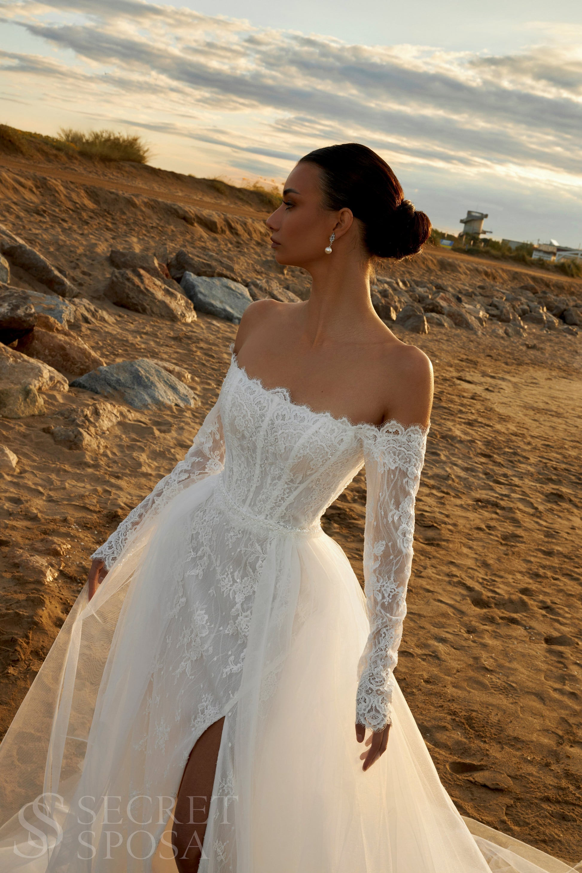 Bride in lace wedding dress with long off-the-shoulder sleeves, slit skirt, detachable tulle overskirt, open boat-shaped back, and sweep court train.
