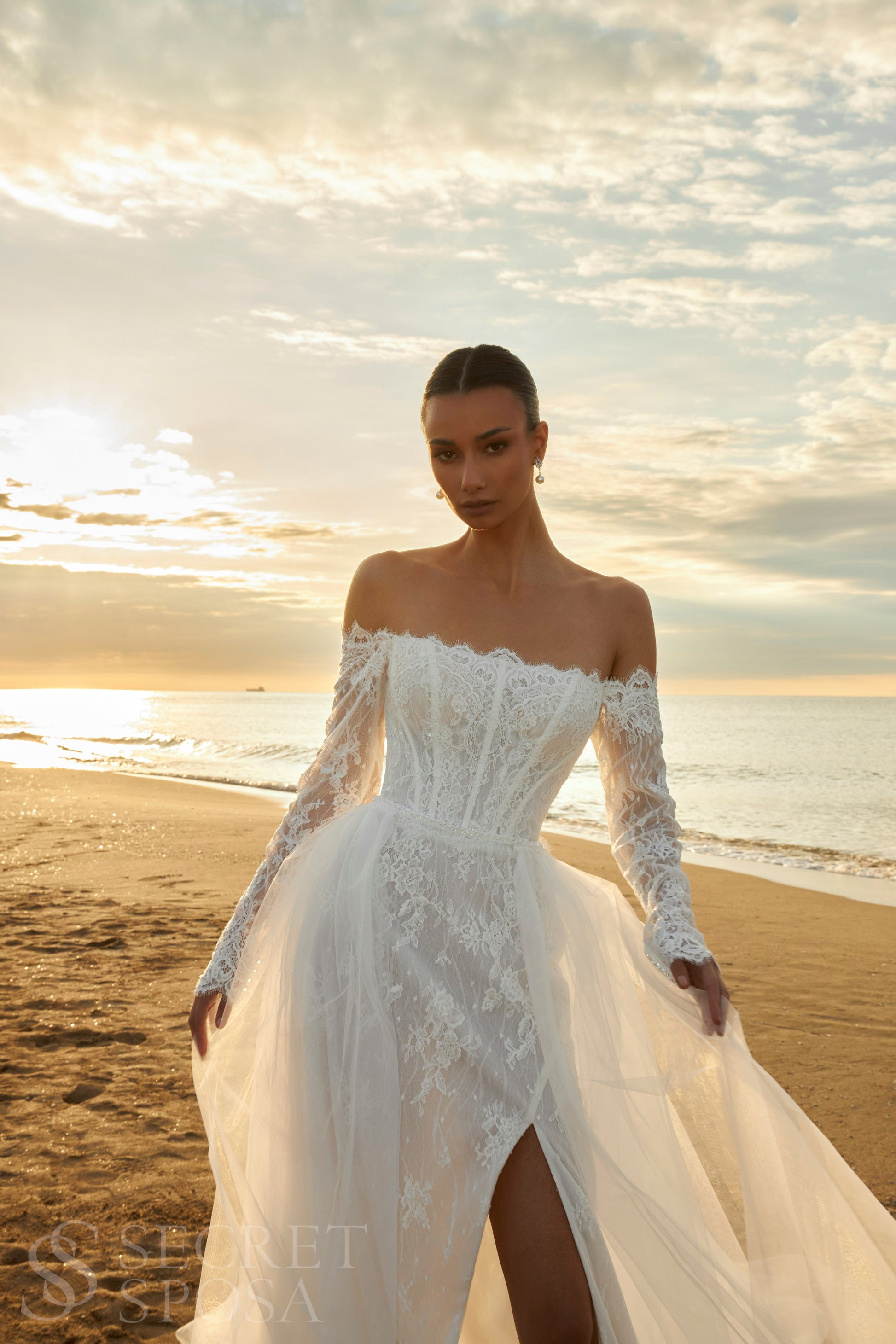Bride in lace wedding dress with long off-the-shoulder sleeves, slit skirt, detachable tulle overskirt, open boat-shaped back, and sweep court train.