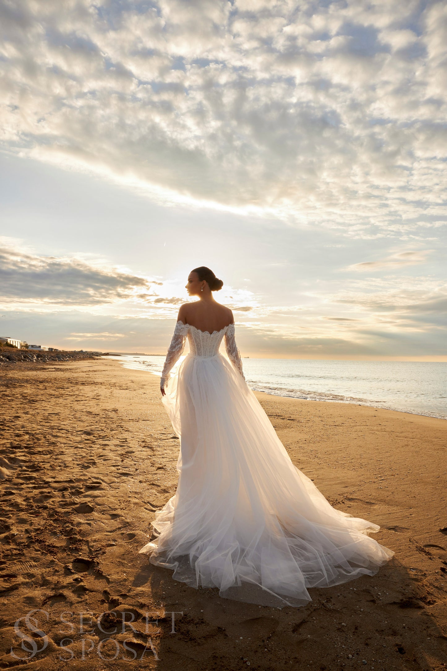 Bride in lace wedding dress with long off-the-shoulder sleeves, slit skirt, detachable tulle overskirt, open boat-shaped back, and sweep court train.