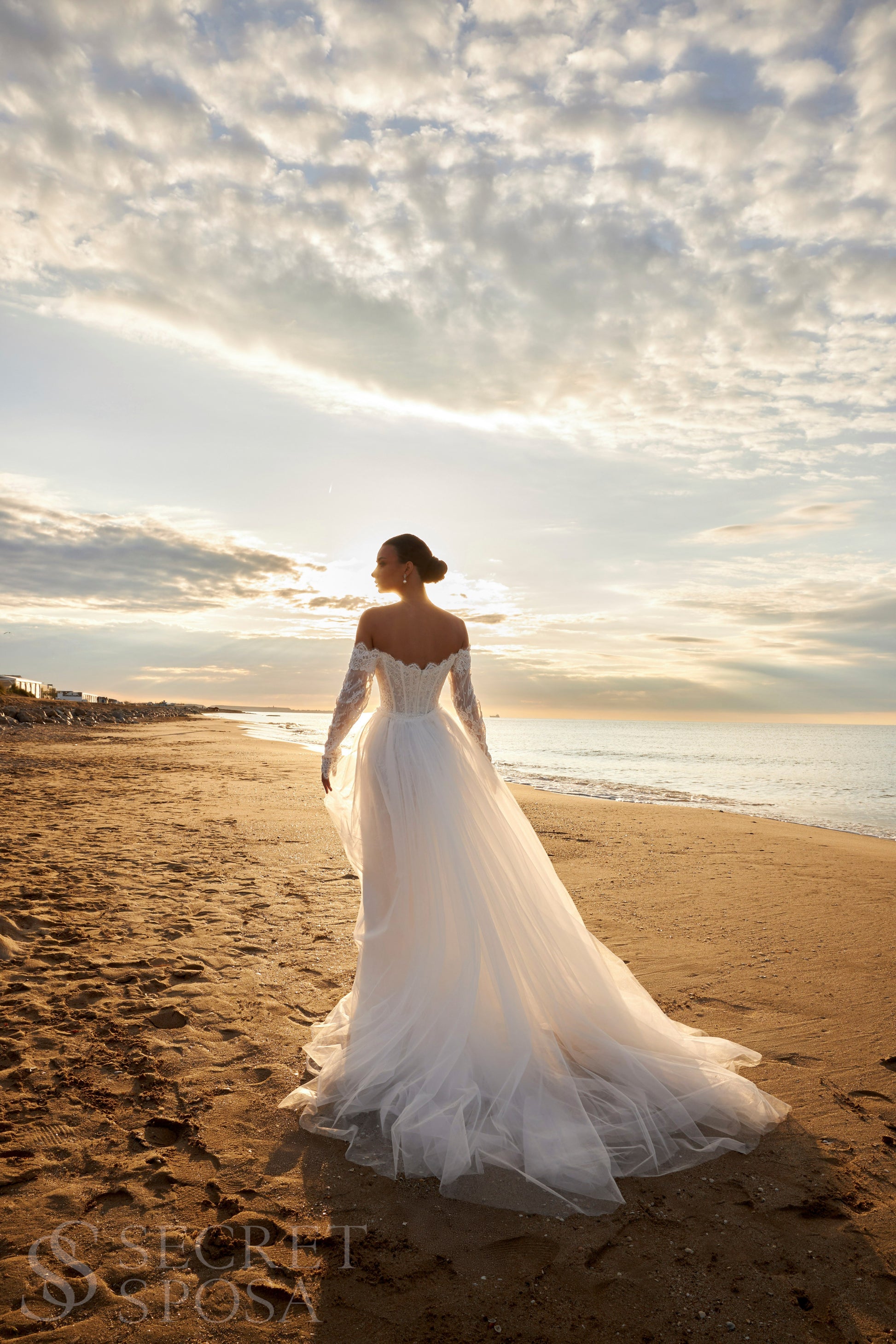 Bride in lace wedding dress with long off-the-shoulder sleeves, slit skirt, detachable tulle overskirt, open boat-shaped back, and sweep court train.