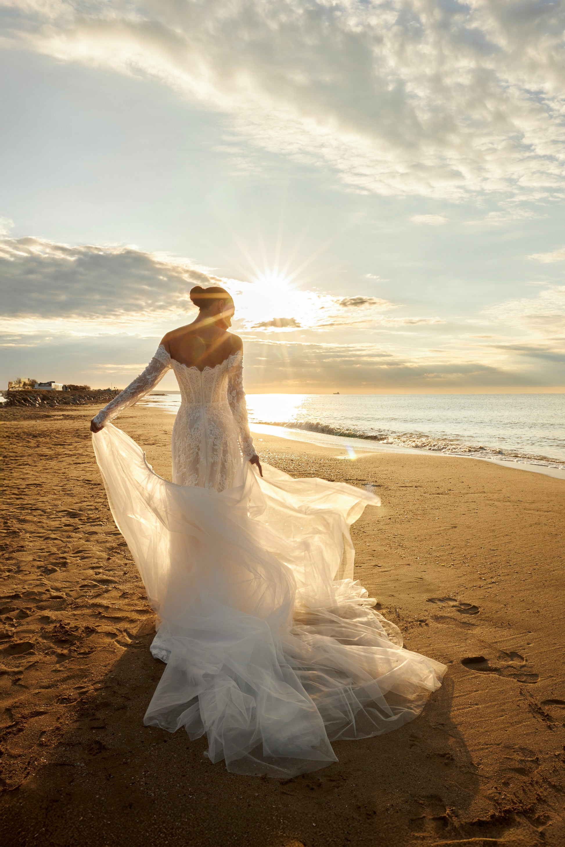 Bride in lace wedding dress with long off-the-shoulder sleeves, slit skirt, detachable tulle overskirt, open boat-shaped back, and sweep court train.