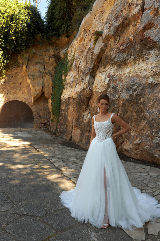 Bride wearing sleeveless A-line wedding gown with boat neckline, pearl and sequin lace bodice, glitter tulle slit skirt, open scoop back, and zip closure.