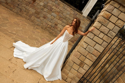 Bride in princess-style sleeveless A-line wedding gown with boat neckline, illusion deep-V pearl and sequin embroidered bodice, satin skirt, and cathedral train.