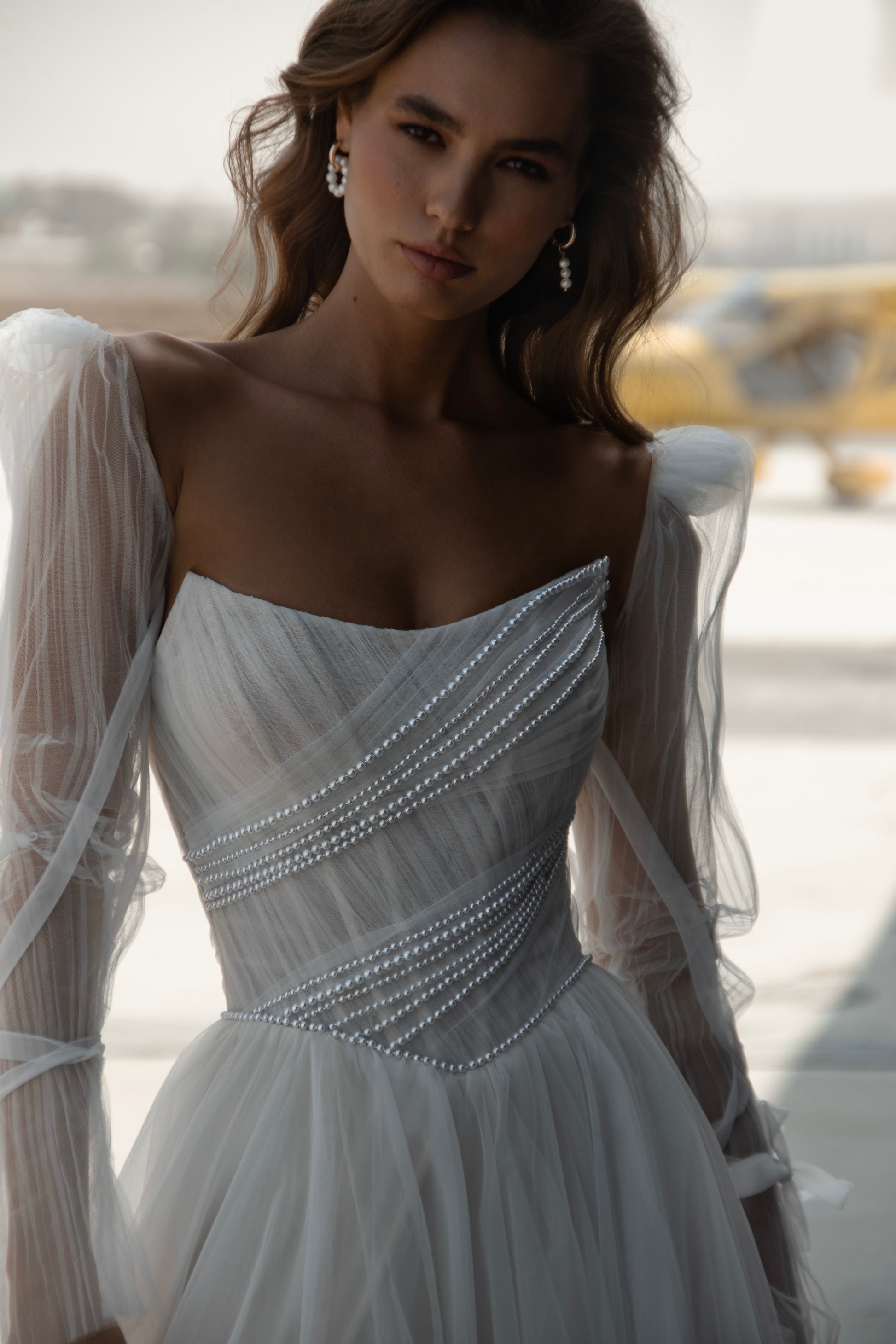 Bride in tulle A-line wedding dress with puff sleeves, pearl-draped corset bodice, and front slit, standing beside an airplane.