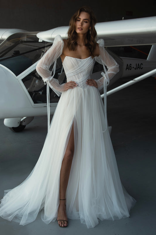 Bride in tulle A-line wedding dress with puff sleeves, pearl-draped corset bodice, and front slit, standing beside an airplane.