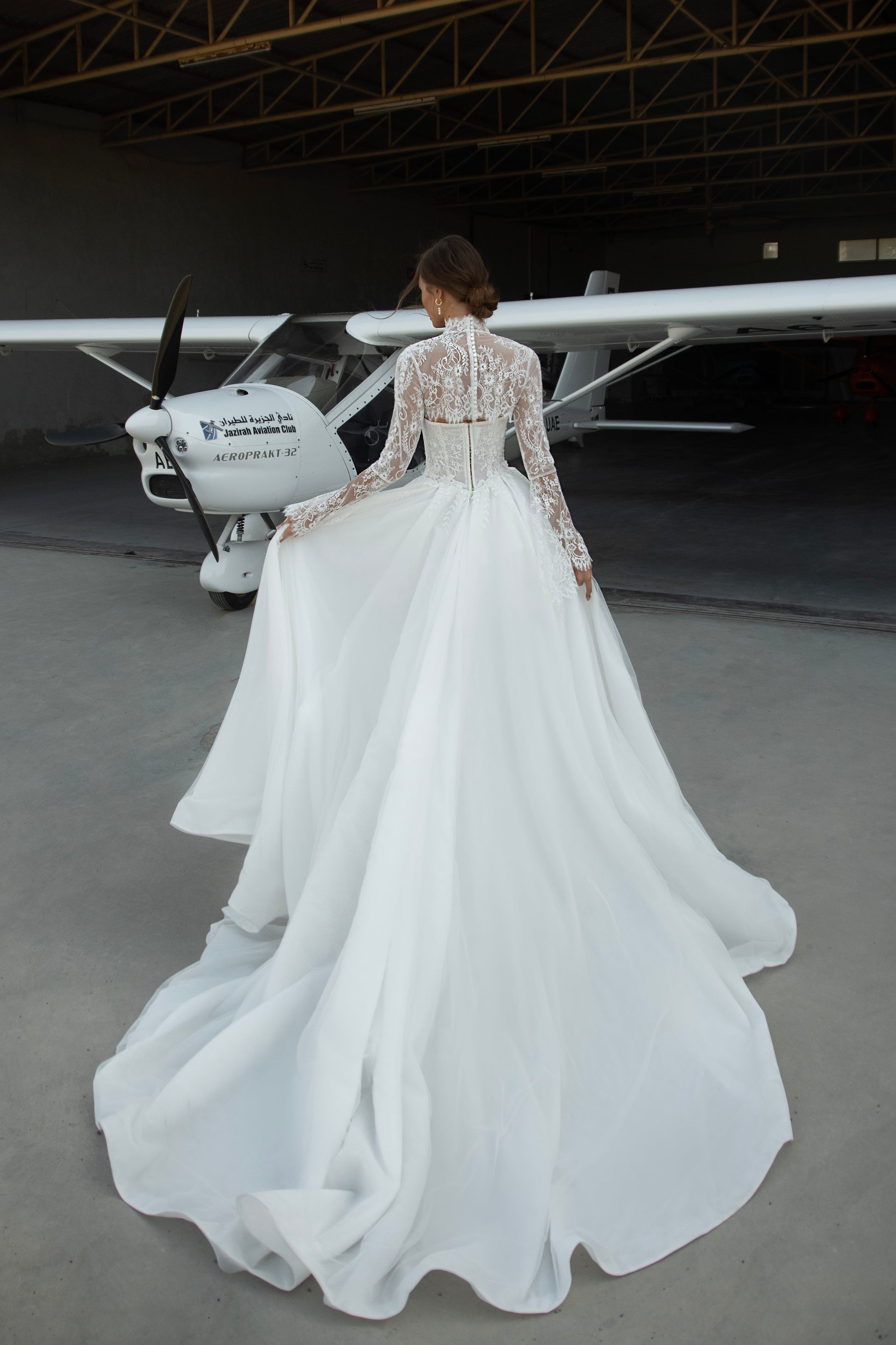 Bride wearing lace long-sleeve wedding dress with high illusion neckline, fitted corset bodice, and chapel train, featuring intricate lace cuffs and back detailing.