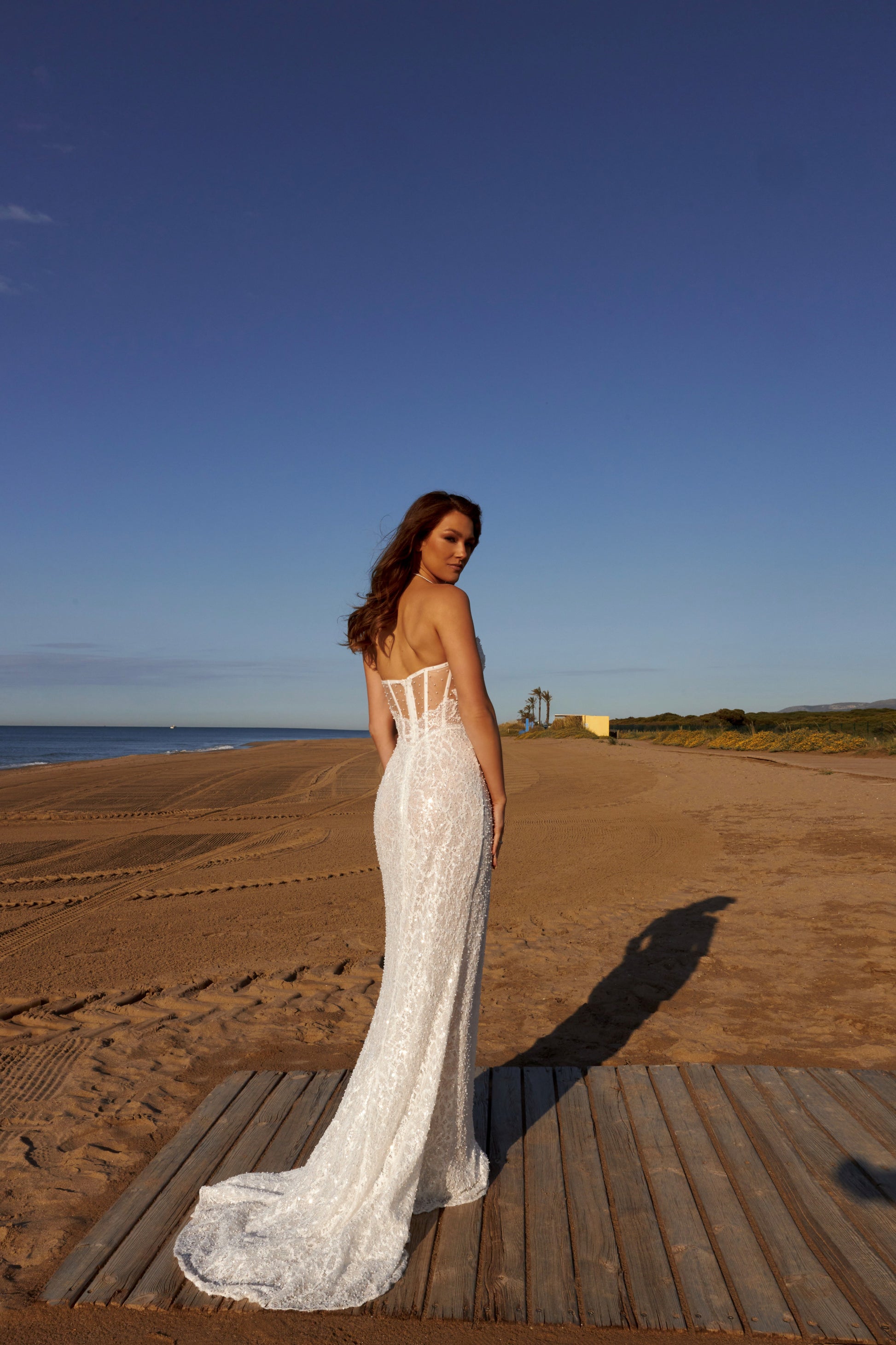 Bride in sleeveless lace bridal gown with sweetheart neckline, pearl and sequin embellishments, drop-down waist, slit skirt, zip closure, and sweep train.