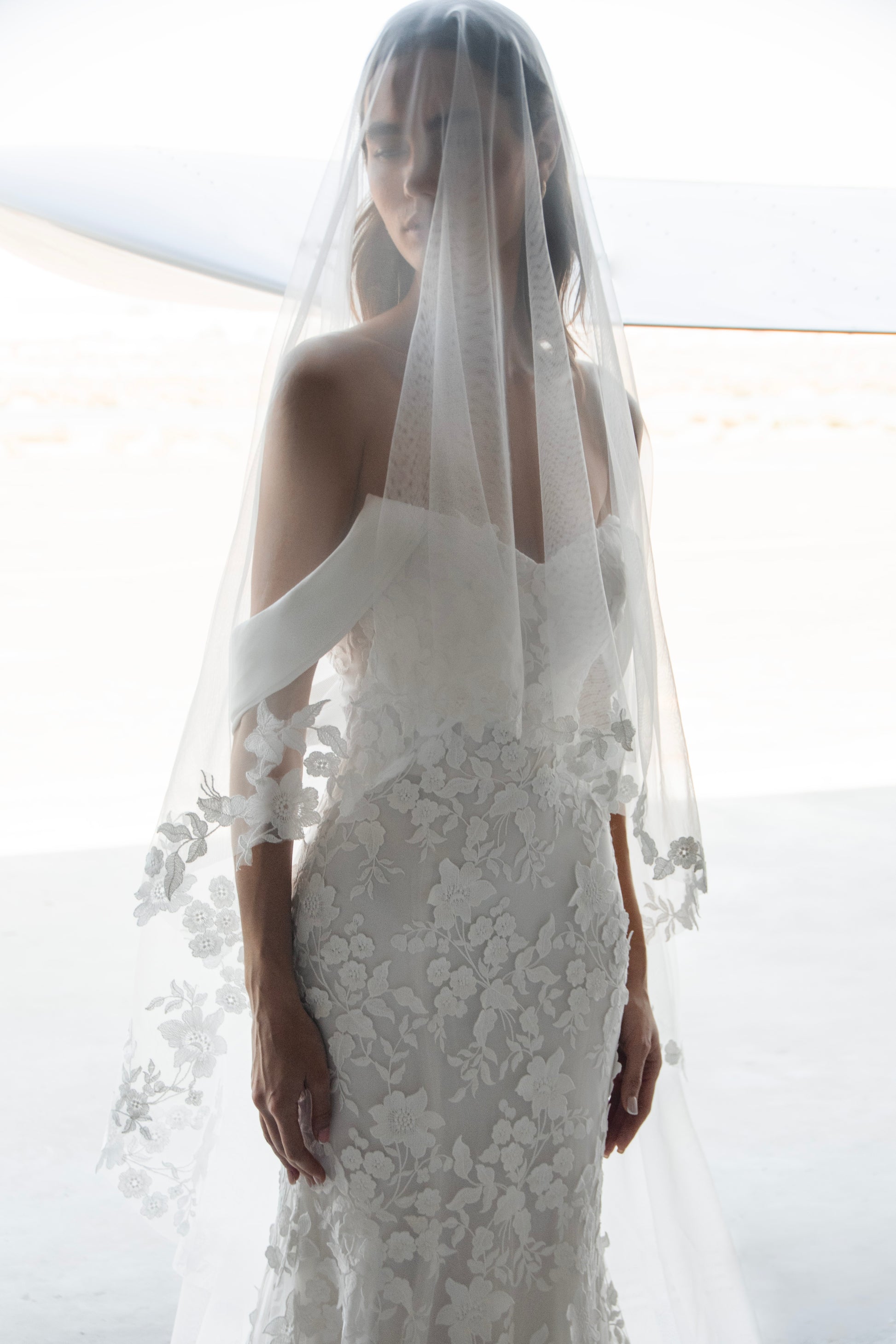 Bride in long-sleeve lace fit-and-flare wedding dress with floral appliqués, illusion back, and chapel train; styled with optional satin off-shoulder wrap.