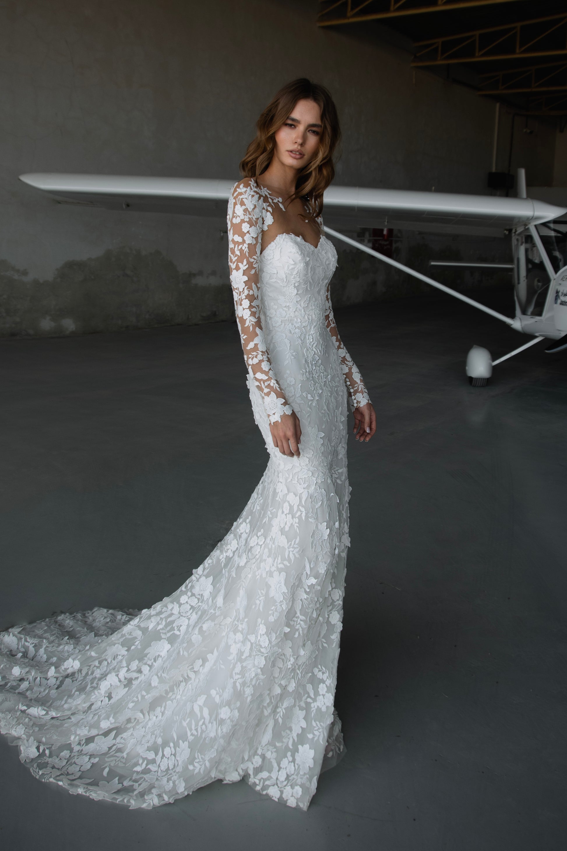Bride in long-sleeve lace fit-and-flare wedding dress with floral appliqués, illusion back, and chapel train; styled with optional satin off-shoulder wrap.
