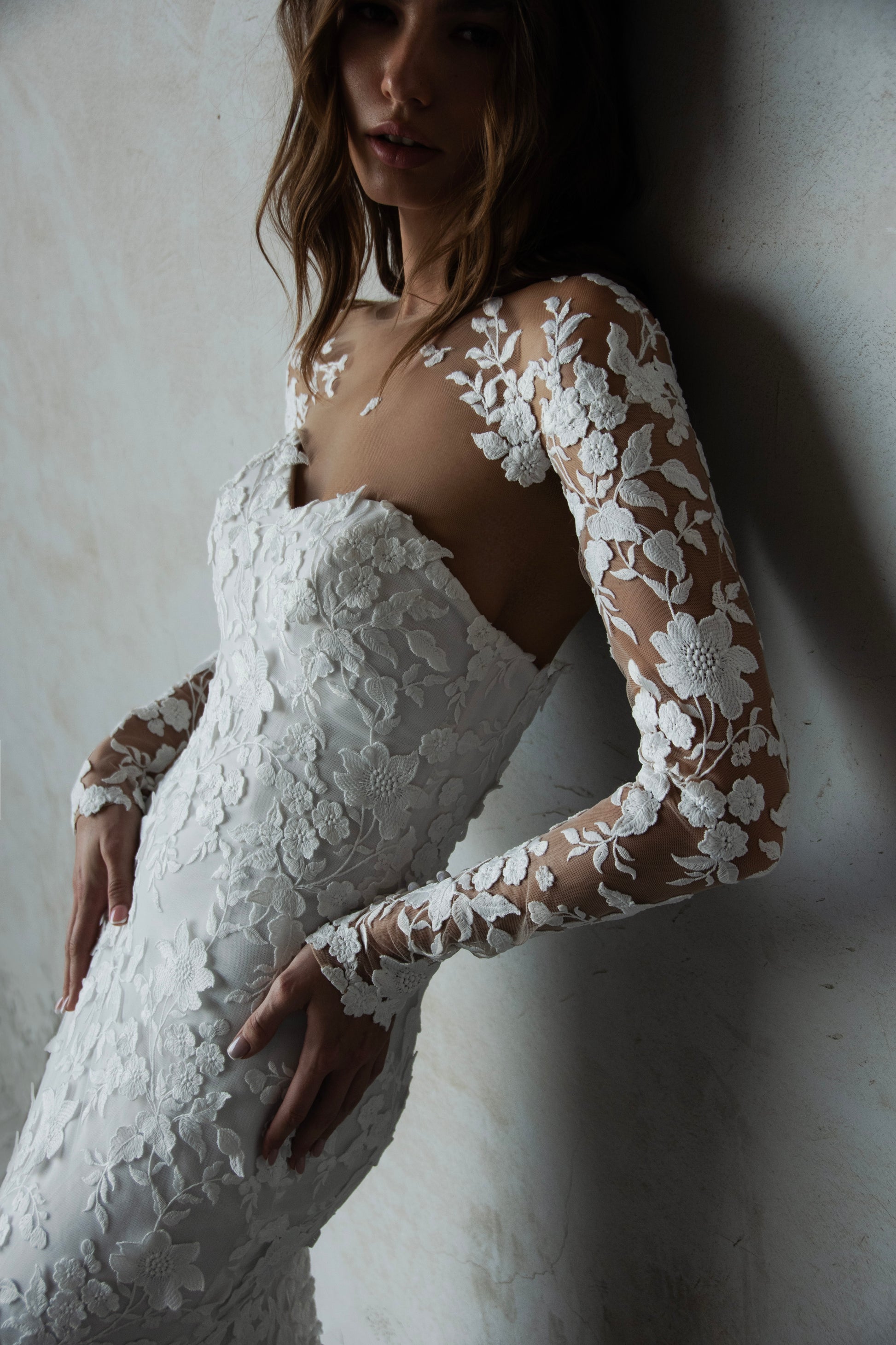 Bride in long-sleeve lace fit-and-flare wedding dress with floral appliqués, illusion back, and chapel train; styled with optional satin off-shoulder wrap.