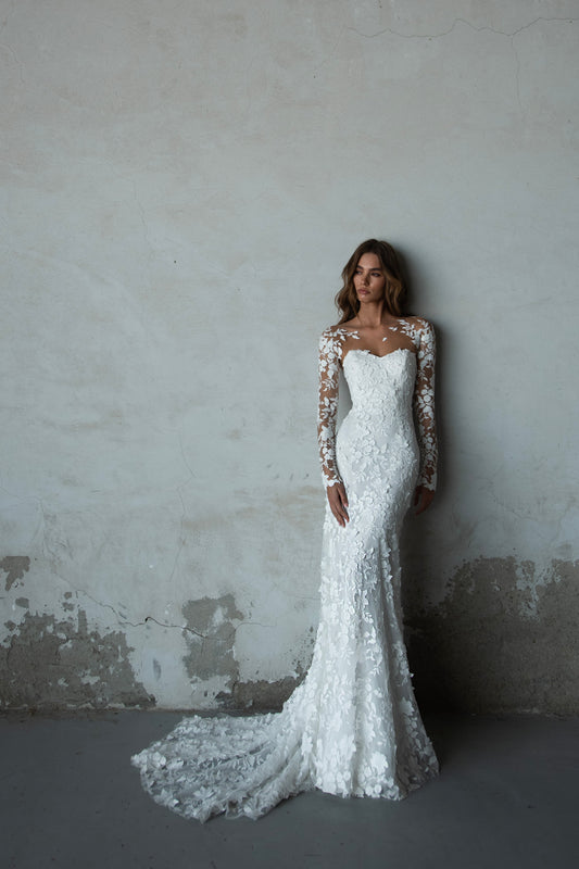 Bride in long-sleeve lace fit-and-flare wedding dress with floral appliqués, illusion back, and chapel train; styled with optional satin off-shoulder wrap.