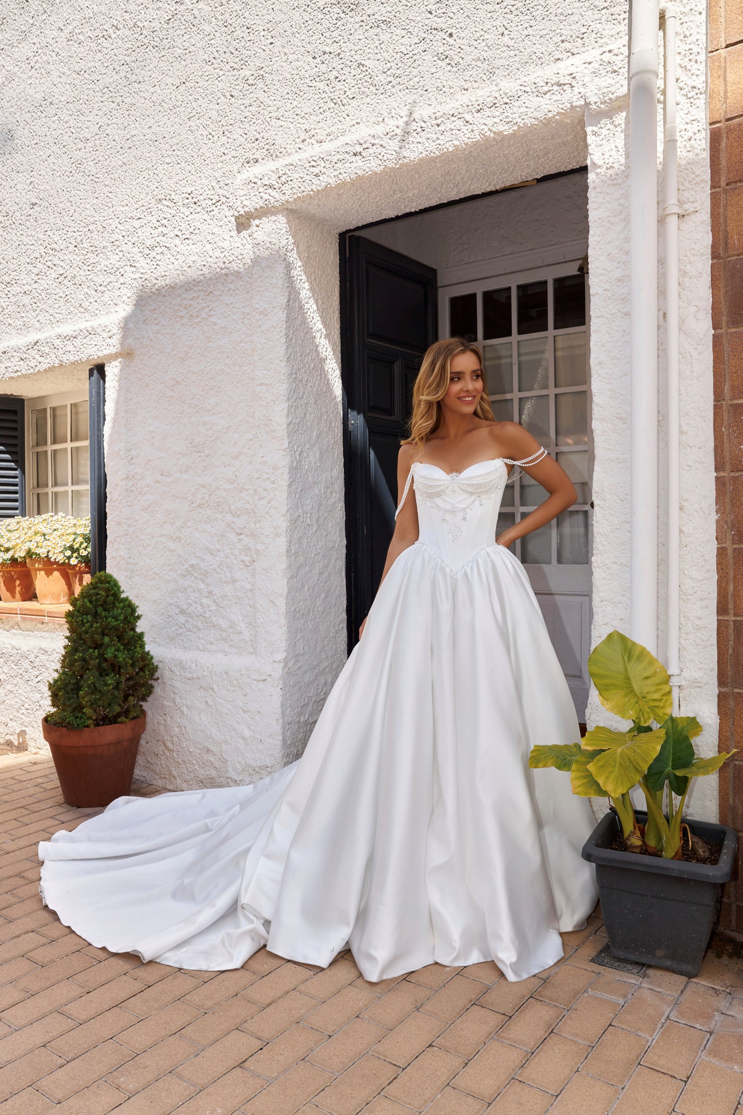 Bride wearing sleeveless satin A-line bridal gown with sweetheart neckline, pearl straps, drop-down waist, zip closure, open back, and chapel train.