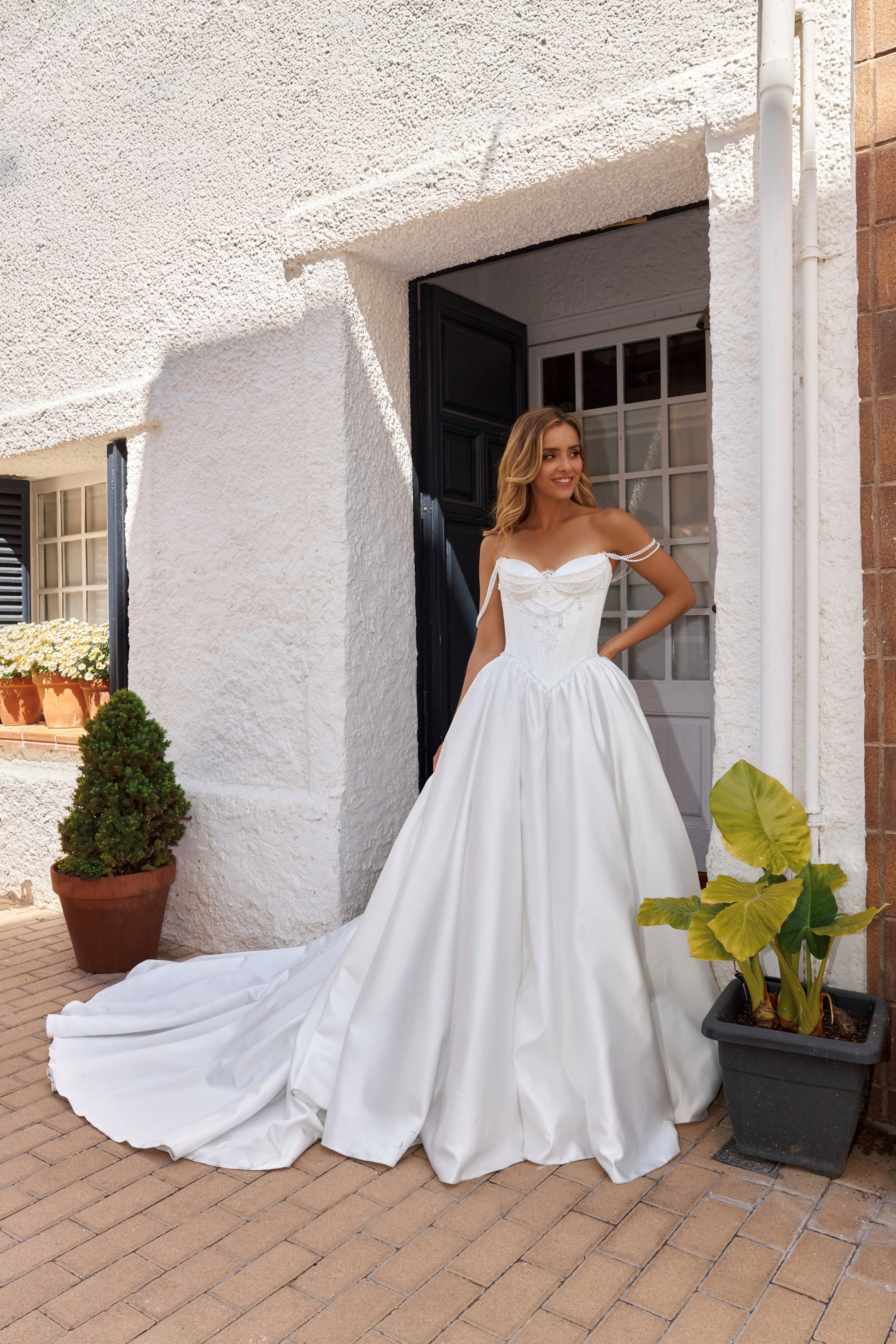 Bride wearing sleeveless satin A-line bridal gown with sweetheart neckline, pearl straps, drop-down waist, zip closure, open back, and chapel train.