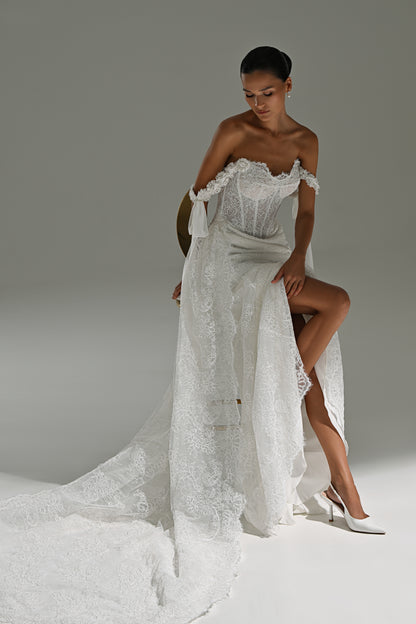 Lace wedding dress with off-shoulder corset, ribbon bows and scalloped train – model seated sideways with the train spread on the floor