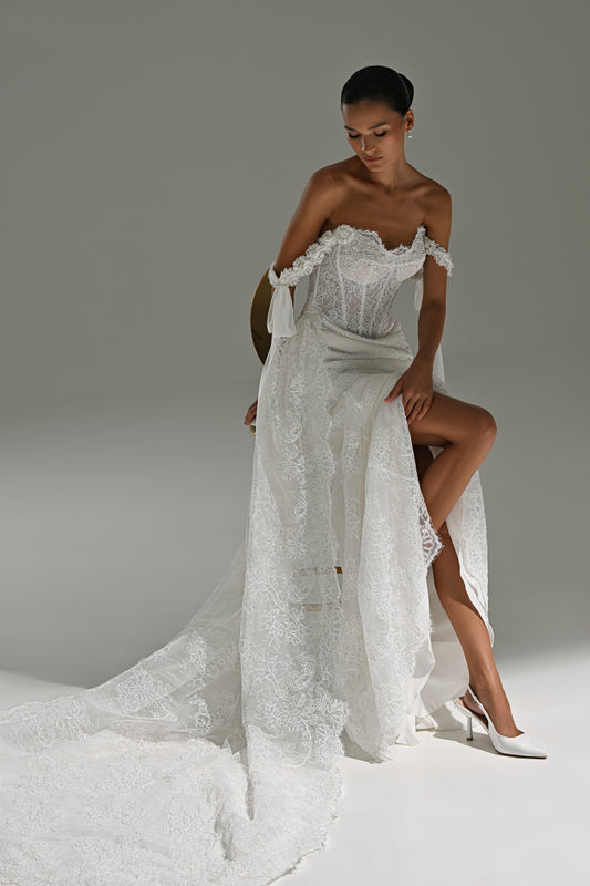 Lace wedding dress with off-shoulder corset, ribbon bows and scalloped train – model seated sideways with the train spread on the floor
