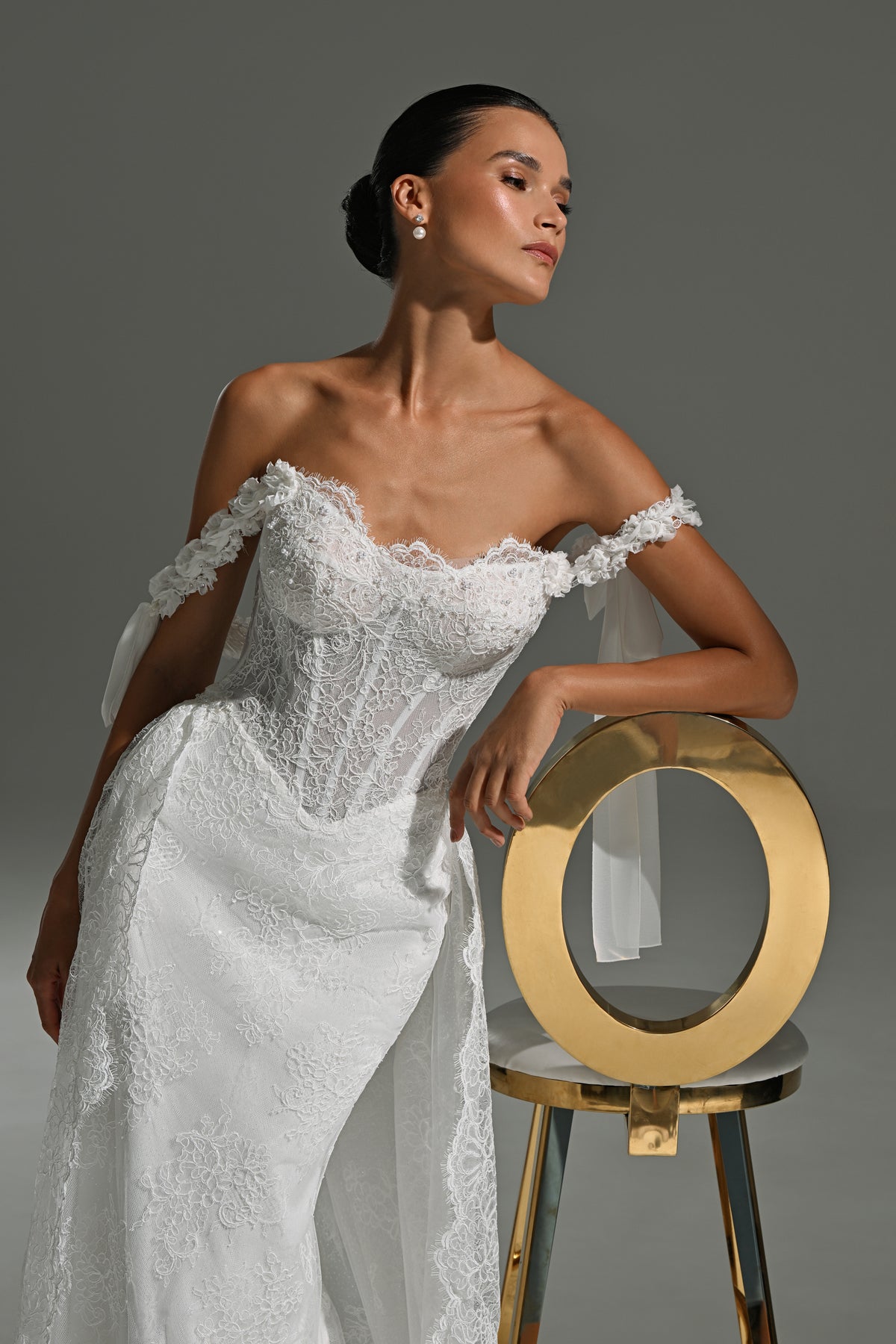 Lace wedding dress with off-shoulder corset and ribbon bows – model leaning on a gold chair, front view of the corset bodice