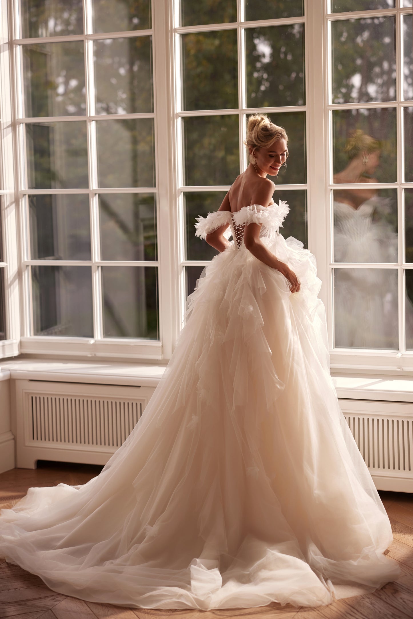 A romantic princess-style tulle wedding gown with a sculpted corset bodice, layered cloud-like skirt, and soft off-shoulder tulle detailing, photographed in natural light by tall windows.