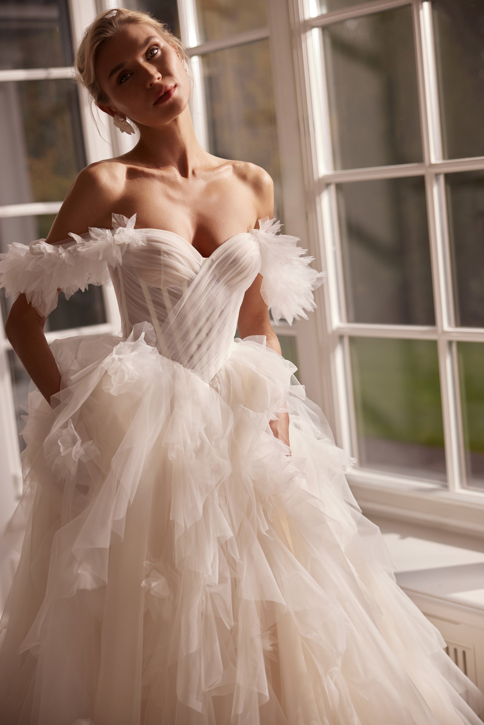 A romantic princess-style tulle wedding gown with a sculpted corset bodice, layered cloud-like skirt, and soft off-shoulder tulle detailing, photographed in natural light by tall windows.