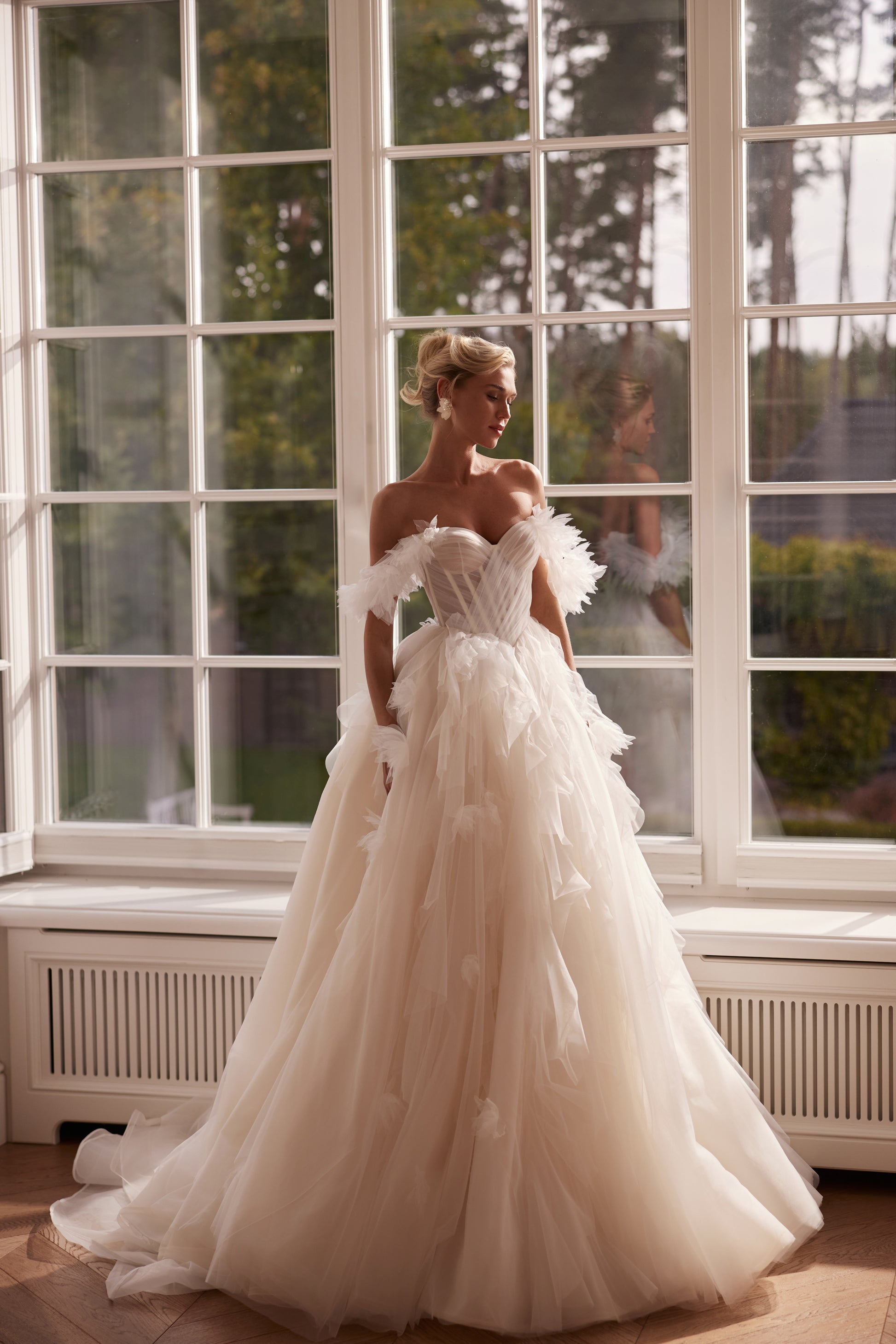 Princess ball gown wedding dress made of layered tulle with an off-shoulder sweetheart corset bodice, feather-textured sleeves, and a full cascading skirt photographed in soft natural light.
