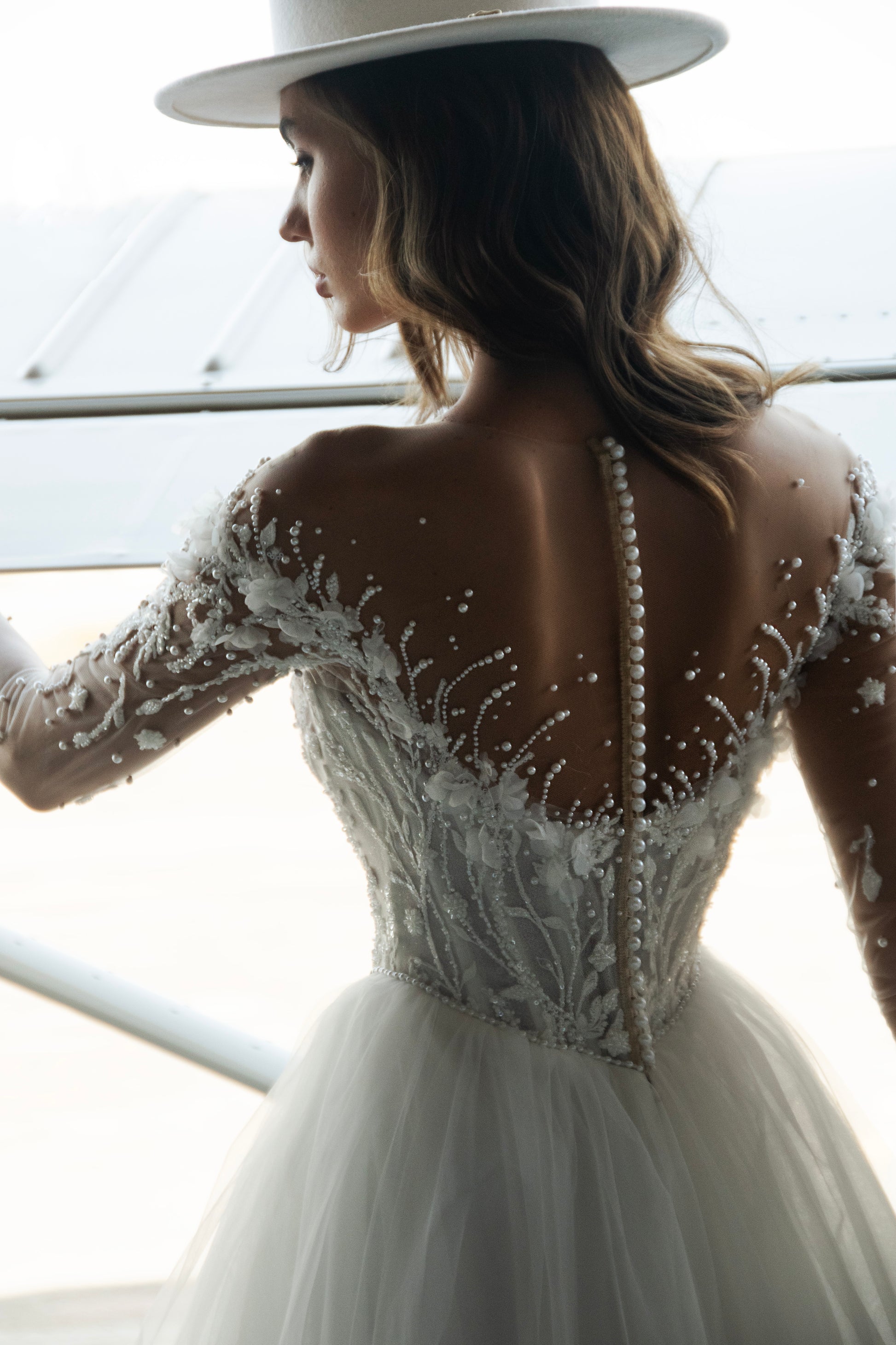 Bride wearing illusion tulle ball gown with 3D floral appliqués, pearl embroidery, and illusion back with buttons, photographed beside an airplane hangar.