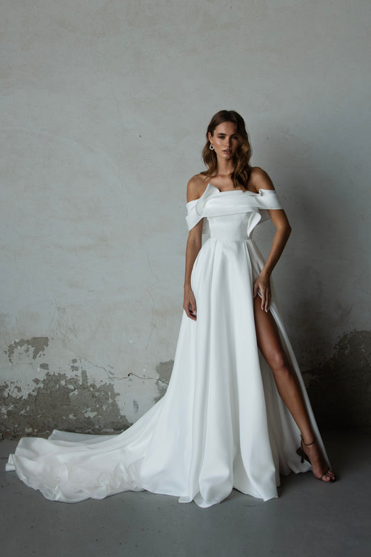 Off-shoulder organza wedding dress with sculpted folded bodice, high-slit skirt, and flowing train in soft white fabric for modern minimalist bridal look