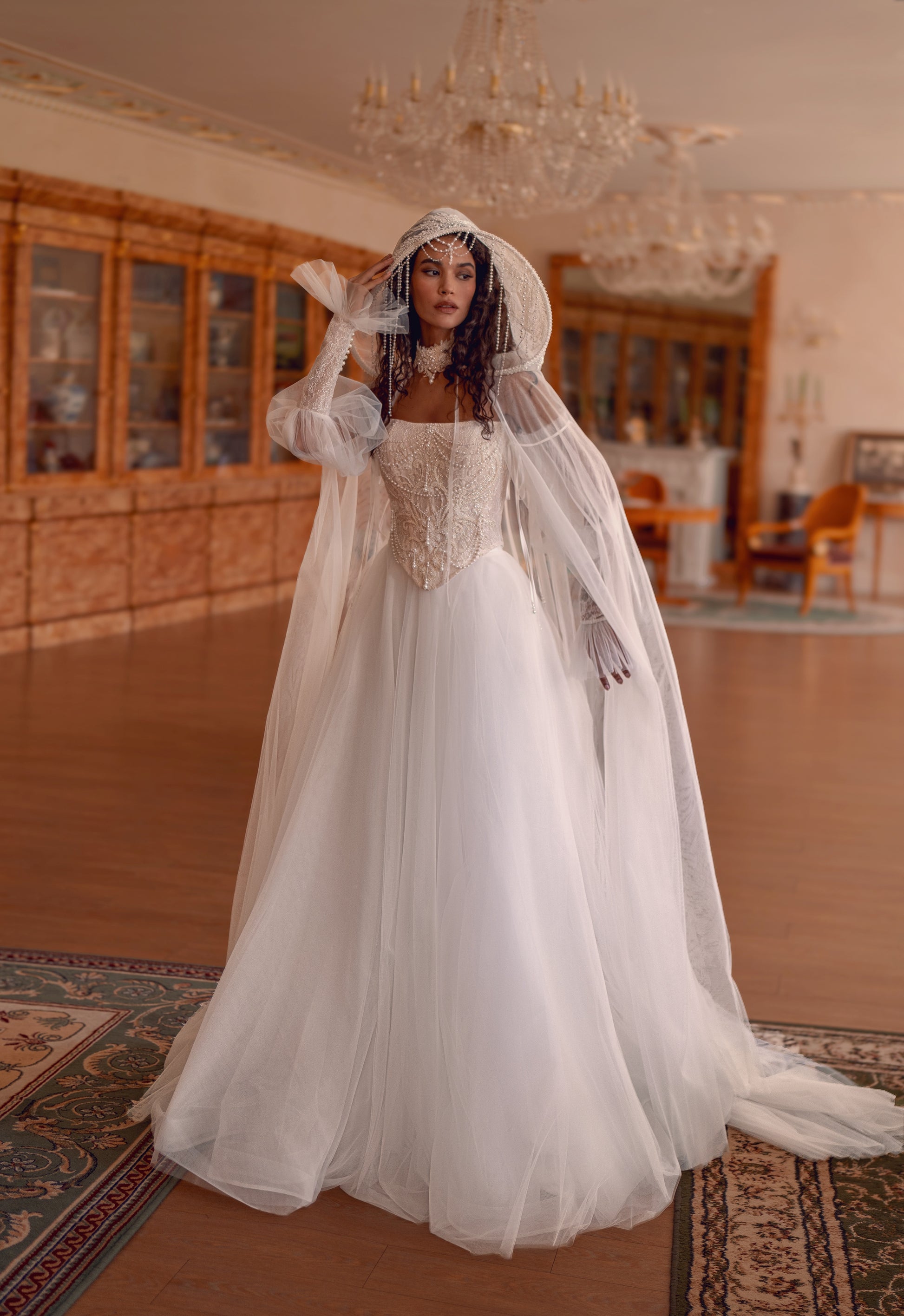 Bride wearing beaded corset A-line wedding dress with detachable tulle sleeves and flowing layered skirt.