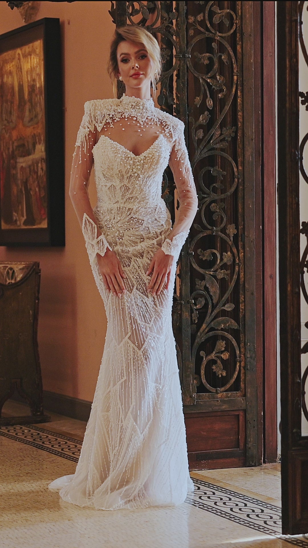 Fitted wedding dress with a sleeveless V-neckline, open back, and detachable cape with long illusion sleeves. Embellished with lace appliques and pearl embroidery, crafted from lace and stretch mesh with a sweep train.