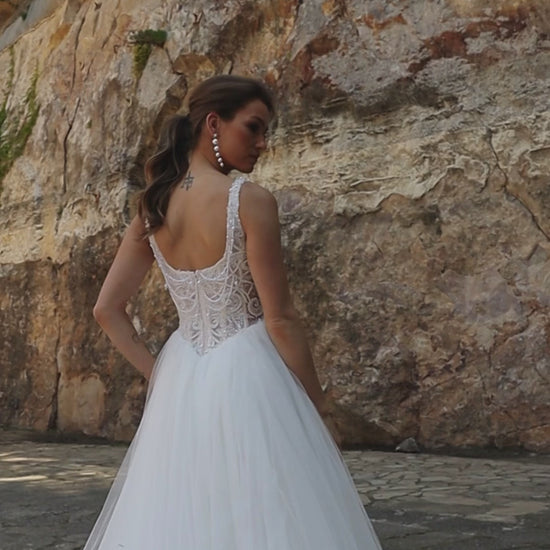 Bride wearing sleeveless A-line wedding gown with boat neckline, pearl and sequin lace bodice, glitter tulle slit skirt, open scoop back, and zip closure.