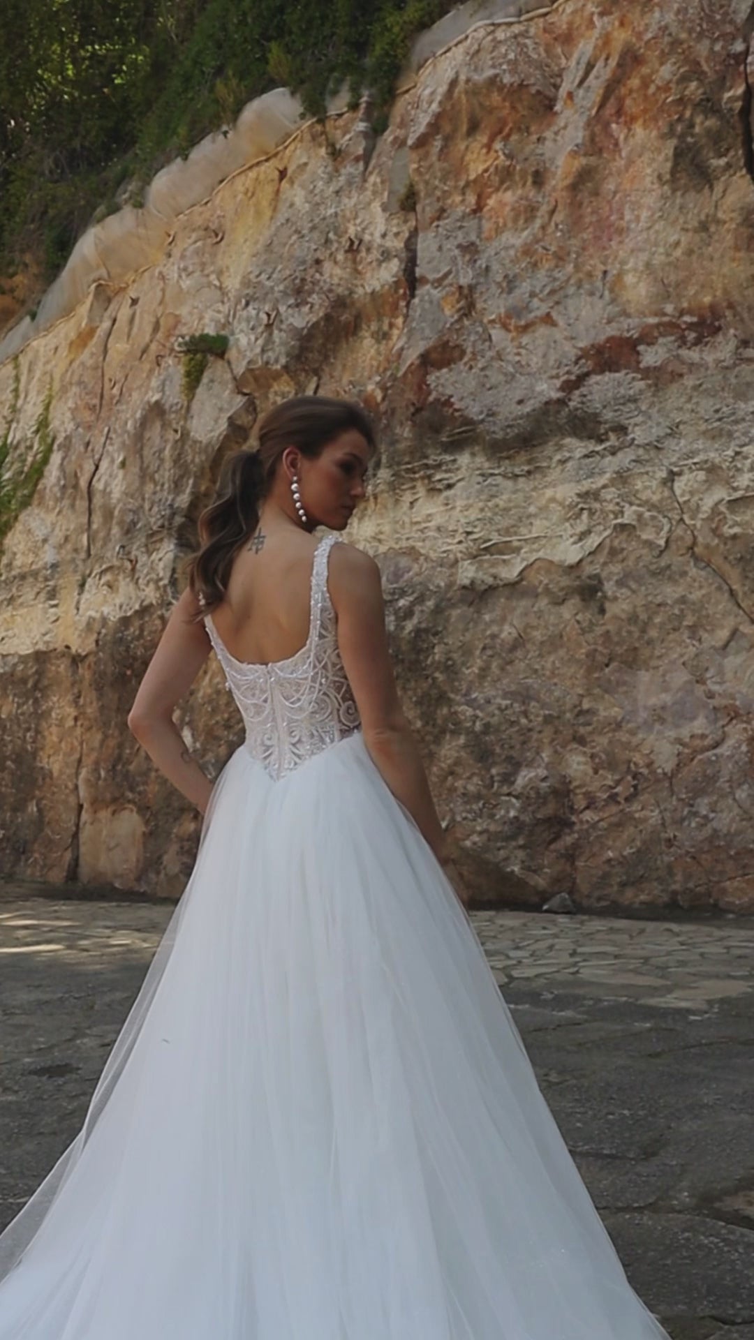 Bride wearing sleeveless A-line wedding gown with boat neckline, pearl and sequin lace bodice, glitter tulle slit skirt, open scoop back, and zip closure.