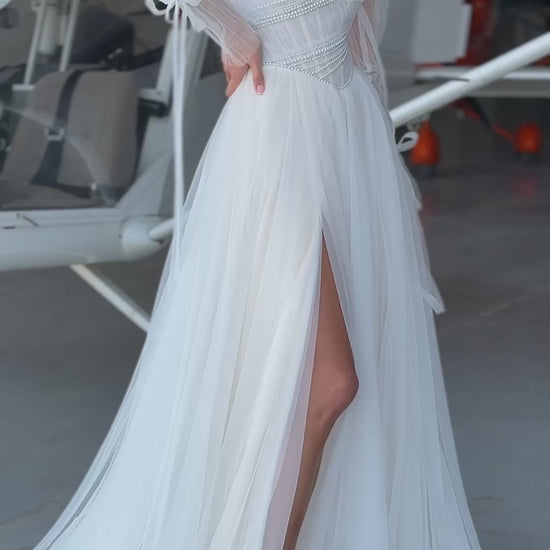 Bride in tulle A-line wedding dress with puff sleeves, pearl-draped corset bodice, and front slit, standing beside an airplane.