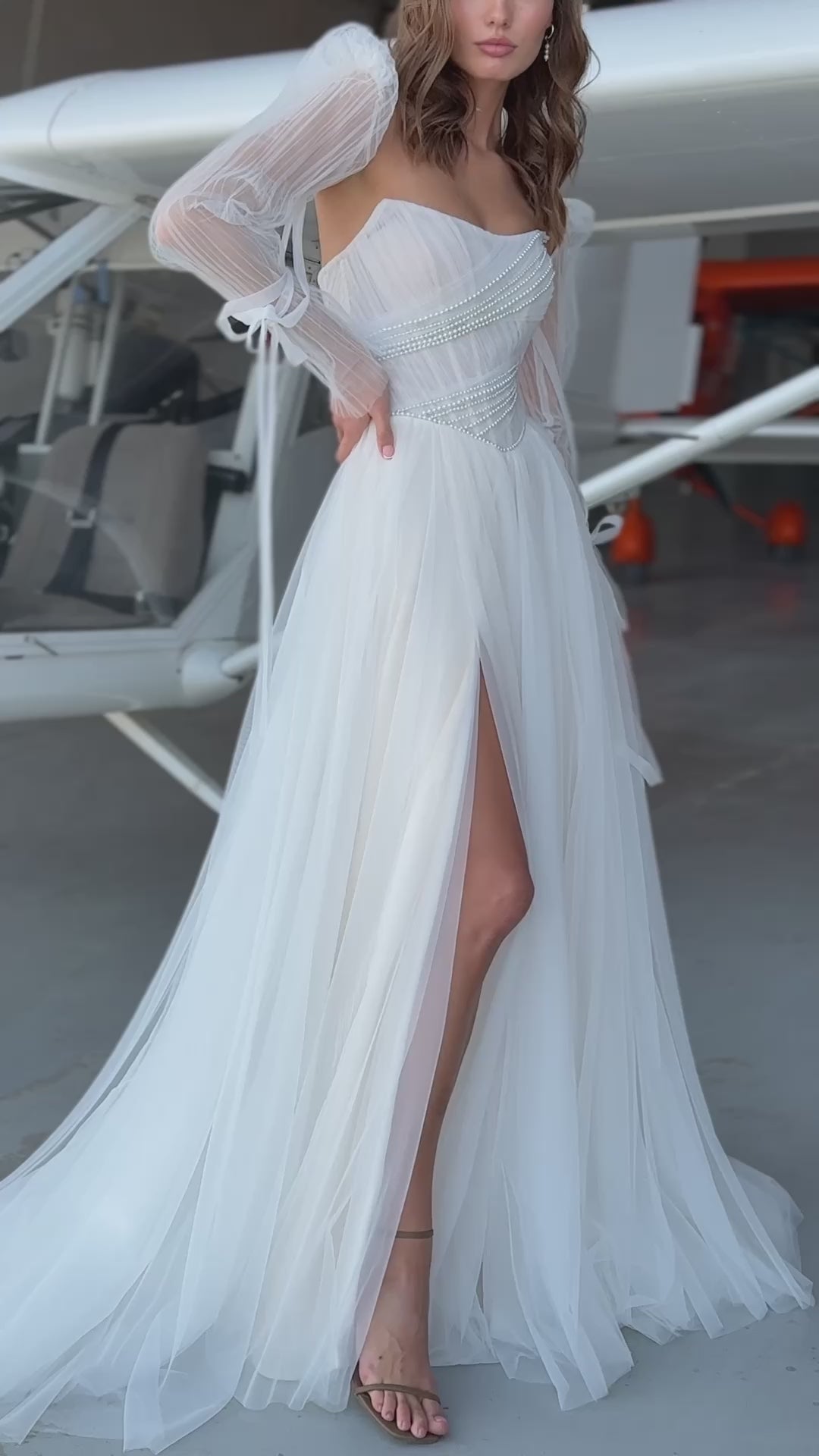 Bride in tulle A-line wedding dress with puff sleeves, pearl-draped corset bodice, and front slit, standing beside an airplane.