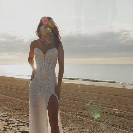 Bride in sleeveless lace bridal gown with sweetheart neckline, pearl and sequin embellishments, drop-down waist, slit skirt, zip closure, and sweep train.