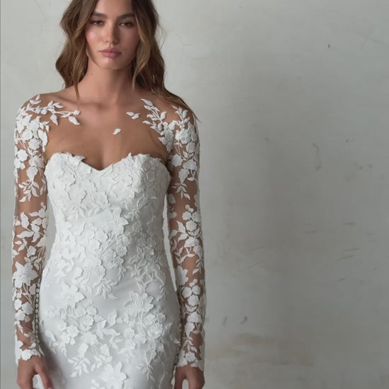 Bride in long-sleeve lace fit-and-flare wedding dress with floral appliqués, illusion back, and chapel train; styled with optional satin off-shoulder wrap.